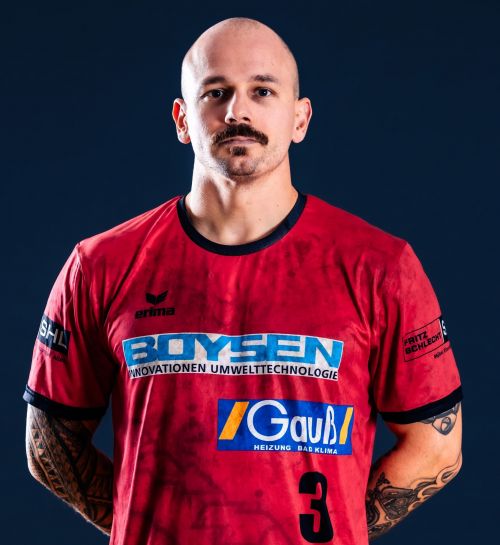 Porträt eines kahlköpfigen Mannes mit Schnurrbart in rotem Sporttrikot mit der Nummer 3 vor dunklem Hintergrund.