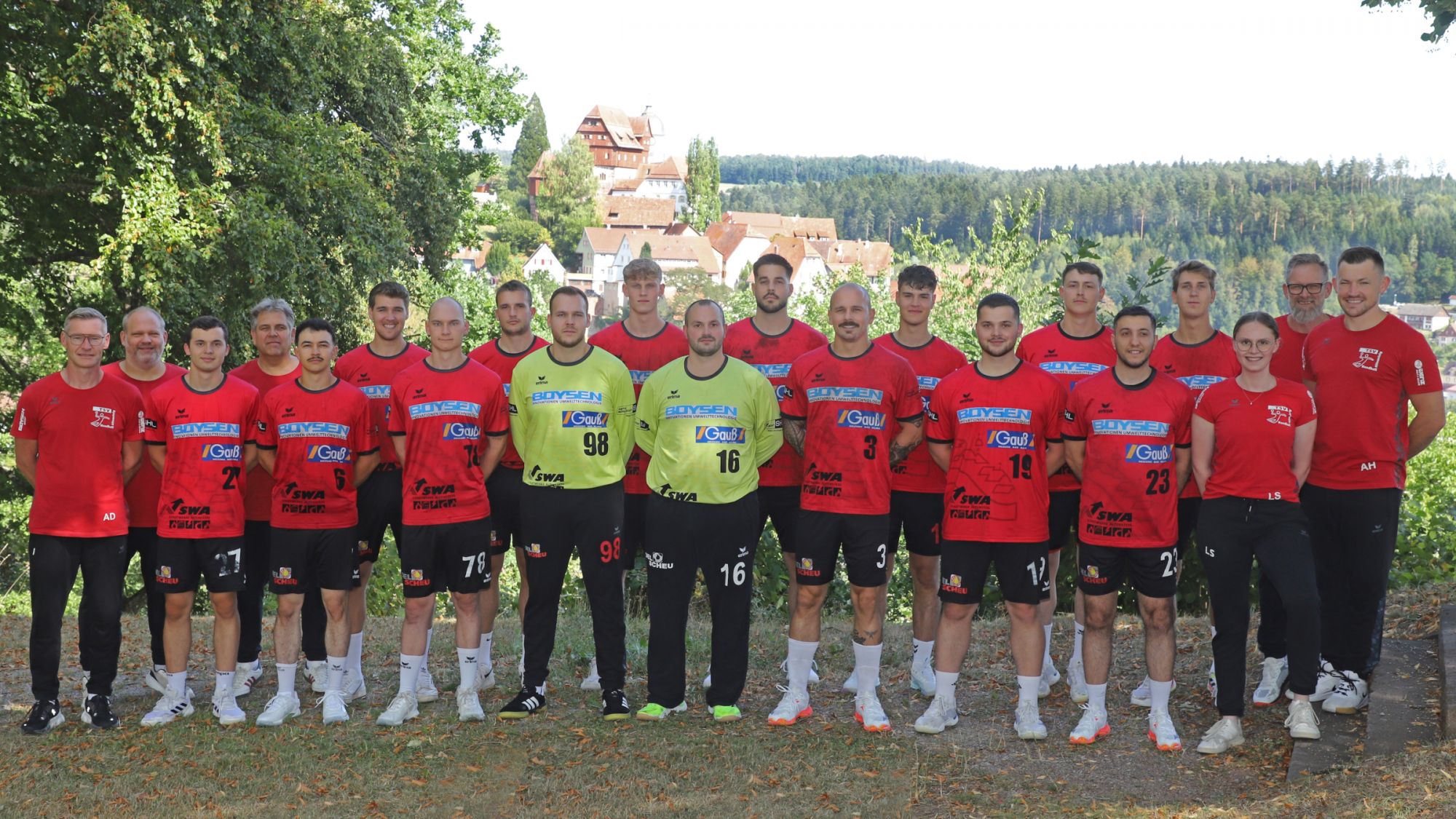 Gruppe von 18 Personen in roten und zwei gelben Fußballtrikots steht im Freien vor Wald und Schloss.