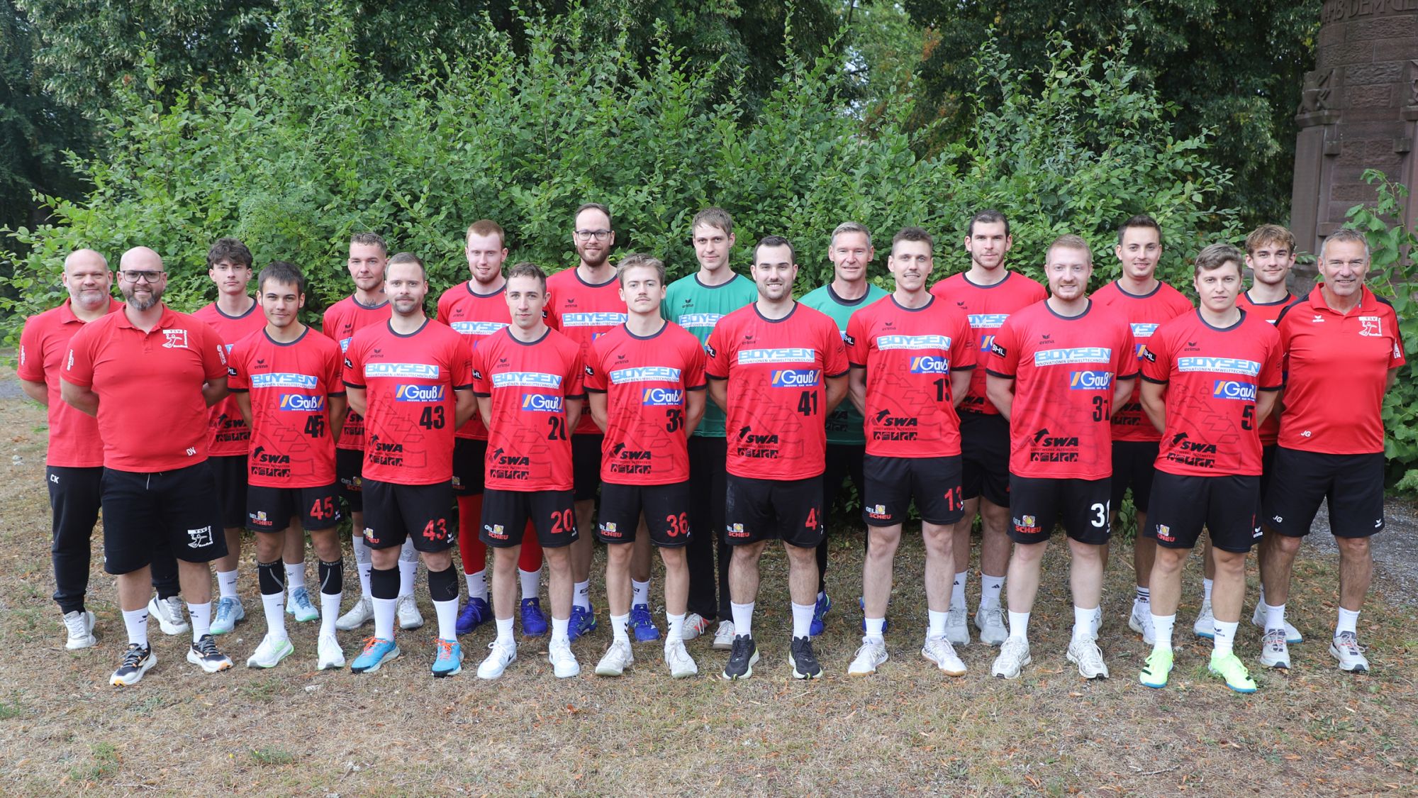 Gruppe von 17 Männern und 2 Kindern in roten Sporttrikots und schwarzen Shorts steht im Freien vor grünem Gebüsch.