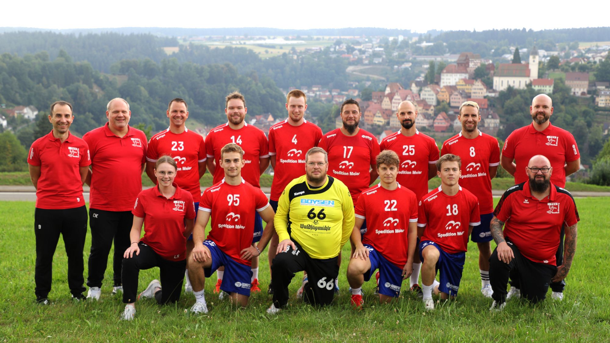 Elf Männer und eine Frau in roten Sportshirts und ein Mann in gelbem Trikot posieren auf einer Wiese vor einer Stadtlandschaft.