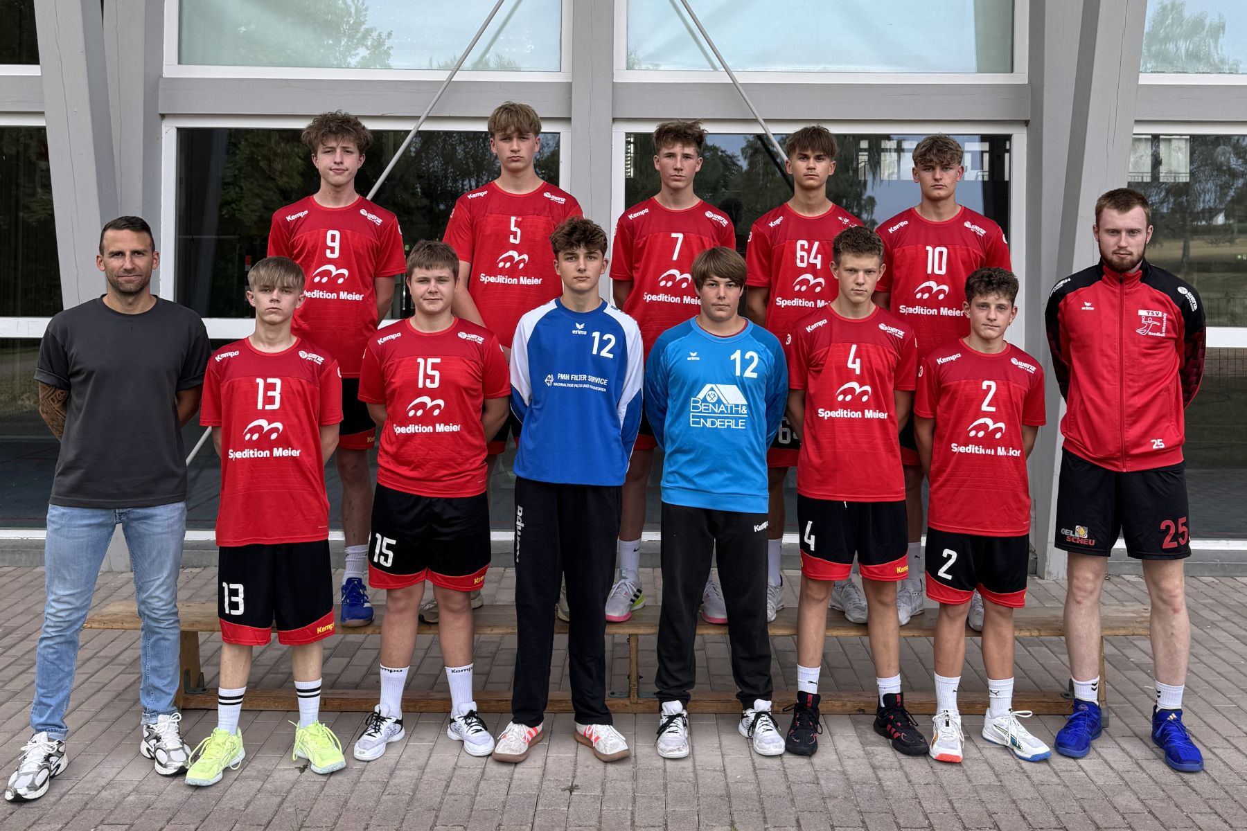 Gruppe von 13 Männern vor einem Gebäude, die meisten in roten Sporttrikots, zwei in blauen Trikots, zwei Männer ohne Trikot.