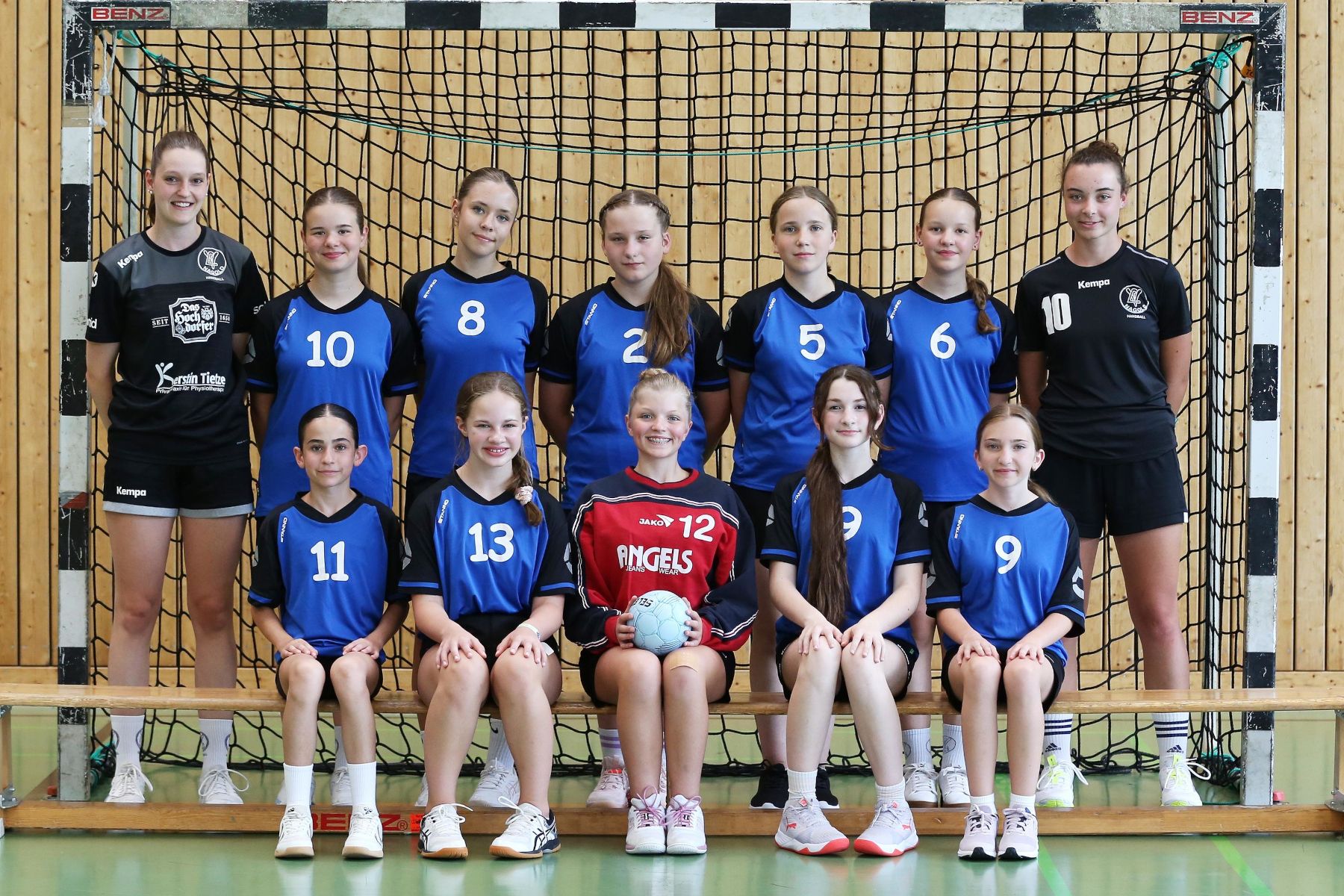 Jugendliche Handballmannschaft mit elf Spielerinnen in blauen und schwarzen Trikots, einige sitzend und einige stehend vor einem Tor.