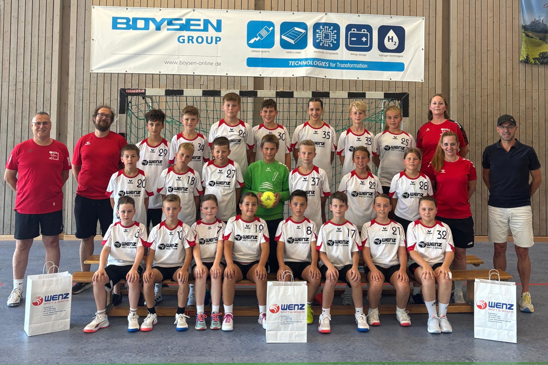 Jugend-Handballmannschaft in drei Reihen mit Trainern vor einer Sponsorenwand; einige halten Geschenktüten, ein Spieler hält einen Ball.