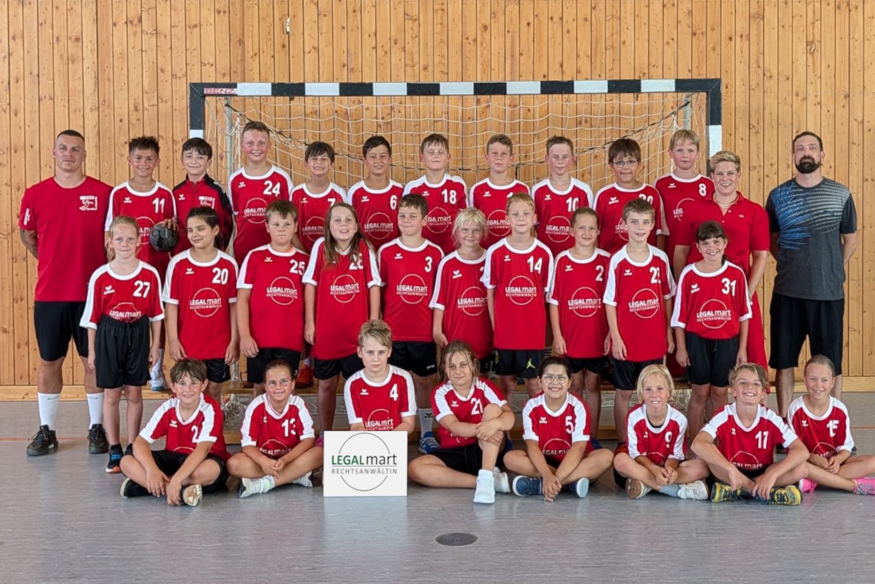 Große Gruppe von Kindern und Erwachsenen in roten Sporttrikots vor einem Handballtor in einer Halle, sitzend und stehend; Schild „LEGALmart“ vorn.