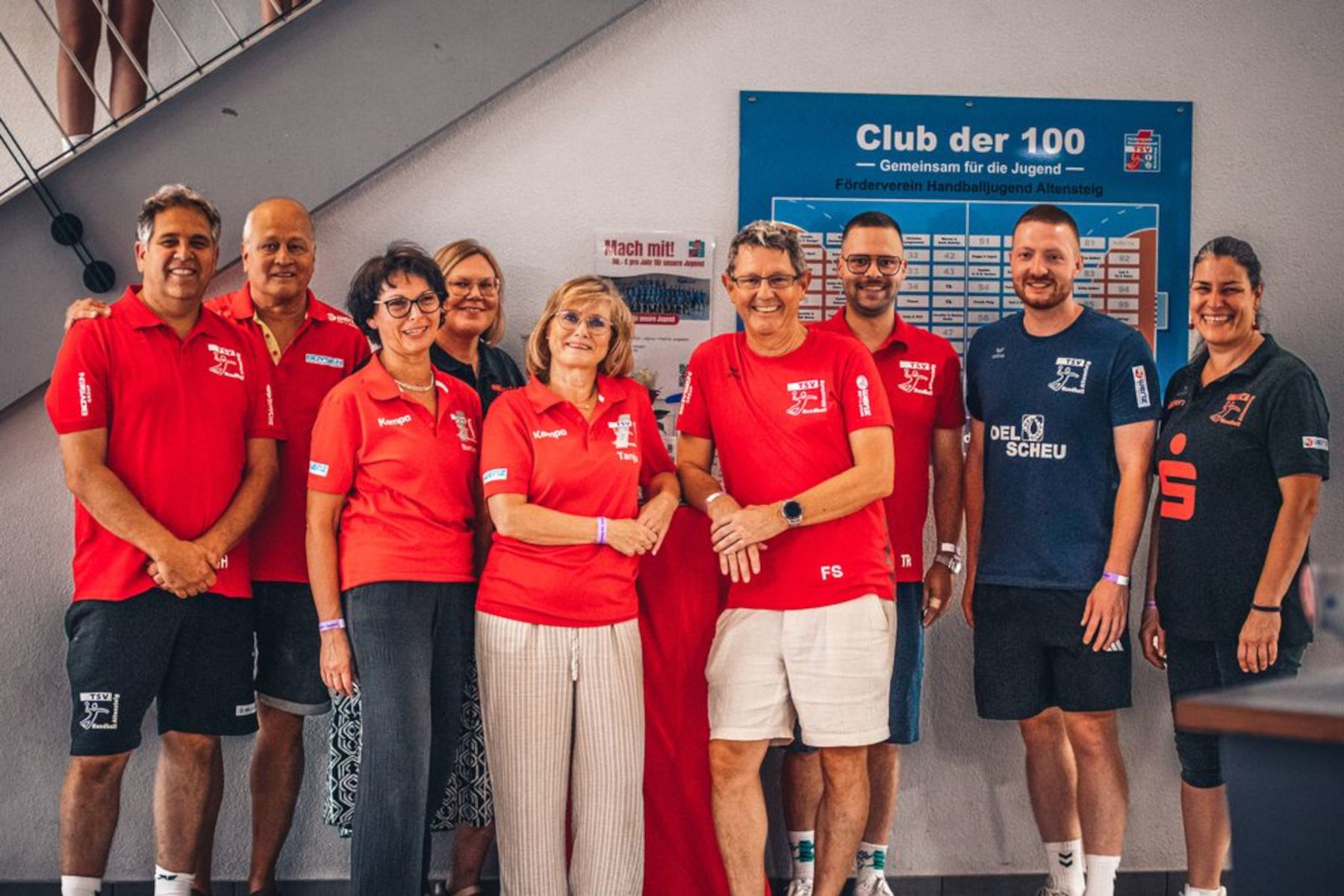 Acht Personen stehen nebeneinander vor einer Wand mit blauem „Club der 100“-Schild.