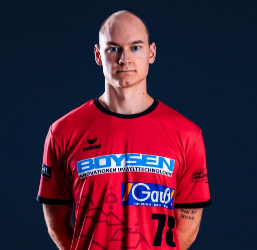 Mann mit rasierter Glatze in rotem Sporttrikot mit mehreren Logos vor dunklem Hintergrund.